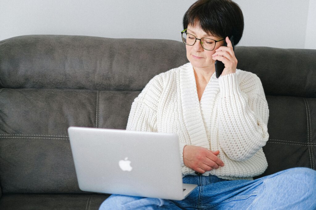 Elderly woman sitting on a sofa, using a laptop and phone simultaneously.